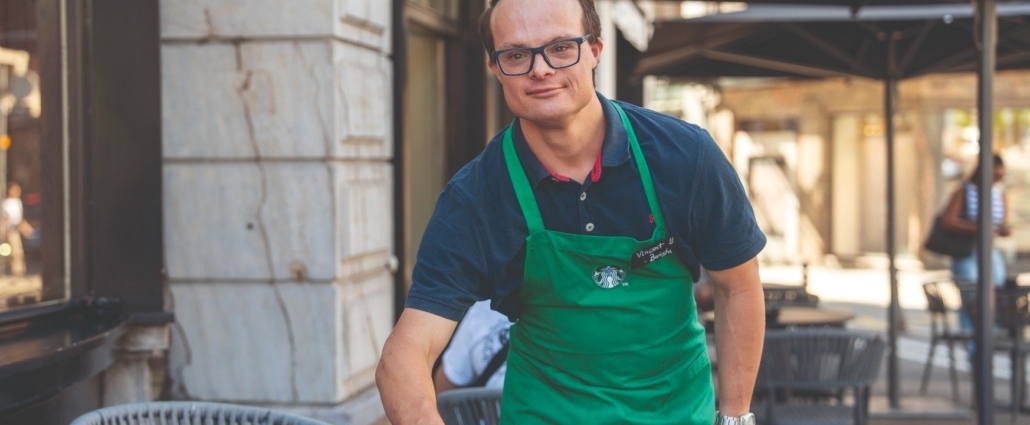 Un homme portant un tablier vert de barista pose devant la terrasse d’un café.